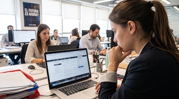 Una mujer frente a una computadora portátil con un sitio de ofertas de empleo abierto, en un entorno de oficina con varias personas.