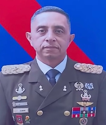 Retrato de medio cuerpo de un militar con uniforme, insignias, condecoraciones y corbata sobre un fondo azul y rojo