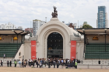 Grupo de personas frente al arco de entrada de un pabellón con tribunas laterales, dos carteles verticales rojos y edificios de fondo