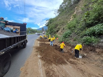 Grupo de trabajadores con camisas amarillas, cascos, palas y picos remueve tierra al costado de una carretera asfaltada. Un camión de carga negro está presente