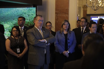 Un hombre con traje y barba cruzado de brazos, una mujer con blazer azul, y varias personas más de pie en un salón con pantalla y araña de luces