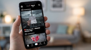 Una mano sostiene un teléfono móvil mostrando la interfaz de TikTok, con un video de una mano con una carta y otro de pétalos que forman un corazón roto.