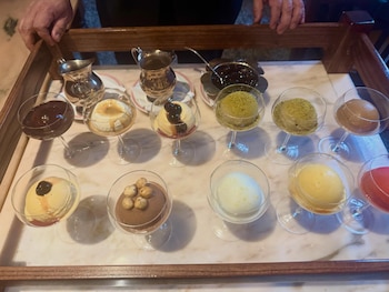 Una persona sostiene una bandeja de madera con diez copas de postres individuales, dos jarras metálicas y un recipiente con salsa oscura