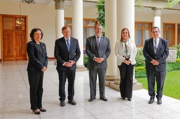 Cinco personas con trajes formales posan de pie en un corredor exterior con columnas blancas frente a un jardín