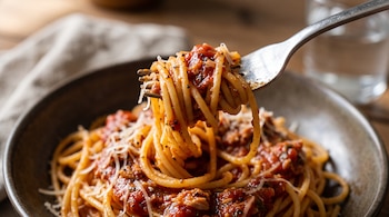 Un tenedor sostiene espaguetis cubiertos de salsa roja, pimienta molida y queso rallado sobre un plato con pasta.