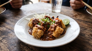 Un plato blanco con trozos de tofu frito y masa frita bañados en salsa, acompañados por col y lima sobre una mesa de madera.