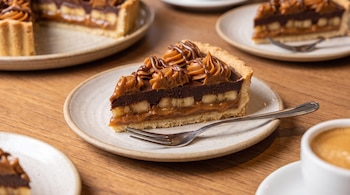 Porción de tarta con capas de dulce de leche, banana y chocolate sobre un plato cerámico con un tenedor pequeño en una mesa de madera.
