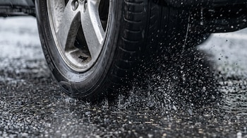 Primer plano de una rueda de coche con neumáticos gastados, salpicando agua sobre asfalto mojado. Los surcos del neumático son apenas visibles.