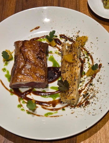 Plato blanco con pieza de carne asada, trozo de col asada, salsas verde y oscura, láminas de cítricos, hojas de perejil y condimentos
