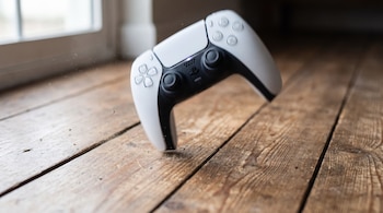 Un mando de PlayStation 5 blanco y negro cayendo hacia un piso de tablas de madera con partículas de polvo, y una ventana borrosa al fondo.