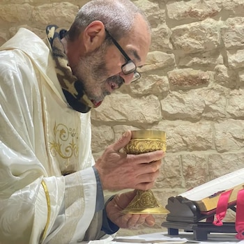 Um padre de túnica branca e óculos segura um cálice dourado. Ao fundo, um livro aberto sobre um cinzeiro e uma parede de pedra. Rosto de perfil em foco