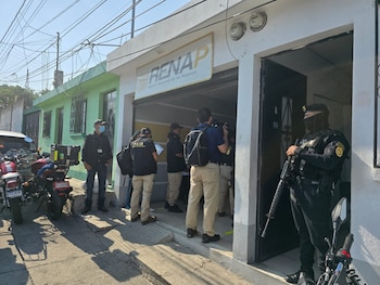 Vista exterior de una oficina de RENAP con varios agentes de policía y personas de pie en la entrada y en la calle, con motocicletas aparcadas