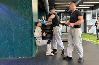 Un hombre hace una sentadilla apoyado en una columna de un gimnasio mientras otros dos hombres apilan discos de pesas sobre sus muslos
