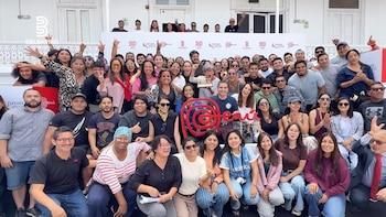 Grupo numeroso de personas sonrientes posa en exteriores, una mujer sujeta un premio, con un logo de Perú grande y un banner que dice "DELBARRIO 20 AÑOS"