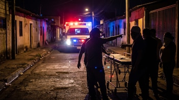 Vista nocturna de una calle oscura donde una ambulancia con luces rojas y azules intermitentes se acerca. Varias personas y una camilla se ven en primer plano.