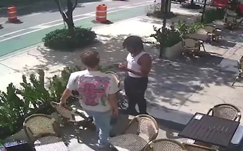 Dos hombres de pie en una acera arbolada con mesas y sillas de café. Hay dos conos de tráfico naranjas en la calle adyacente