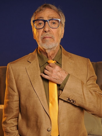 Close de um homem com barba grisalha, óculos azuis, blazer marrom e camisa verde oliva, ajustando uma gravata amarela vibrante sobre um fundo azul escuro