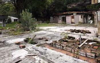 Suelo de concreto agrietado con pilas de ladrillos, restos de muros dañados, árboles al fondo y una persona a la distancia