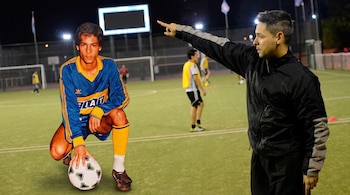 Un hombre de chaqueta negra señala el recorte de un joven futbolista arrodillado con un balón sobre un campo de césped sintético.