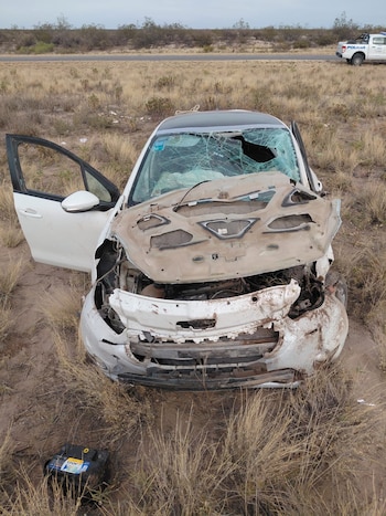 Coche blanco Peugeot gravemente dañado con parabrisas roto y capó abierto, en la maleza junto a una carretera. Una batería está en el suelo y un coche policial se ve a lo lejos
