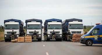 Cuatro camiones blancos y azules, agentes uniformados, cajas y troncos de madera, una camioneta oficial con logo brasileño en una ruta fronteriza con vegetación baja.