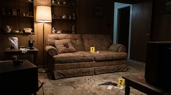 Un sofá con manchas de sangre, un revólver en el piso y marcadores amarillos de evidencia dentro de una sala.