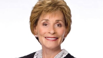 Primer plano de Judy Sheindlin sonriente con toga judicial negra y cuello de encaje blanco sobre fondo blanco