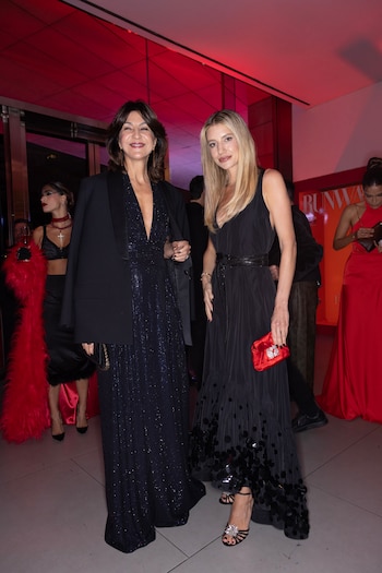 Dos mujeres sonríen en un evento. La de la izquierda viste un vestido azul brillante y blazer negro; la de la derecha, vestido negro con lentejuelas y bolso rojo