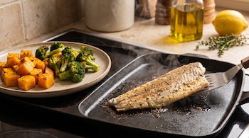 Un filete de merluza en una plancha con espátula, junto a un plato con trozos de calabaza y ramilletes de brócoli. Se ven aceite, limón y tomillo.