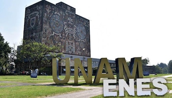 Edificio de la Biblioteca Central de la UNAM con murales y jardines, frente a letras gigantes con las siglas UNAM y la palabra ENES