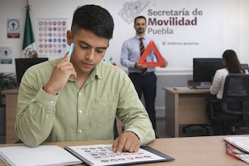 Joven estudia señales de tránsito en un manual. Detrás, un hombre sostiene un triángulo de seguridad. Al fondo, bandera de México y logo Secretaría de Movilidad Puebla. Mujer trabaja.
