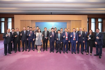 Un grupo de aproximadamente veinte personas vestidas con trajes posa de pie en un salón. La mayoría levanta los pulgares. Se ve una pantalla al fondo