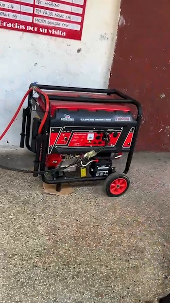 Generador de gasolina rojo y negro con ruedas, una manguera y una batería. Sobre piso de concreto, junto a una pared blanca con un cartel rojo
