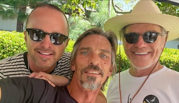 Tres hombres posan sonrientes de cerca; dos llevan gafas de sol y el de la derecha usa un sombrero de paja y una camiseta blanca