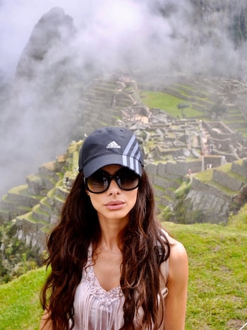 Retrato de una mujer con cabello largo, gorra deportiva negra y gafas de sol grandes, de pie frente a las terrazas verdes y edificios de piedra de Machu Picchu bajo un cielo nublado