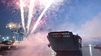 Buque gris en el agua con insignia circular roja y blanca en su popa, fuegos artificiales rojos y blancos en el cielo oscuro, humo, grúas portuarias iluminadas