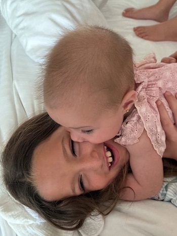 Primer plano de una niña sonriente con cabello castaño, ojos cerrados, y un bebé en un vestido rosa que le besa la mejilla