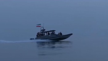 Embarcación gris oscura con bandera iraní en la popa, con varias personas a bordo, navegando sobre agua tranquila y oscura dejando una estela blanca