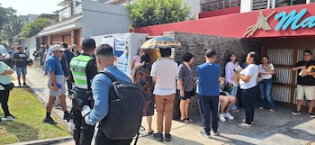 Una multitud de personas espera en una acera bajo el sol, cerca de un edificio con un cartel de votación. Un hombre lleva un chaleco que dice 'SAN BORJA'