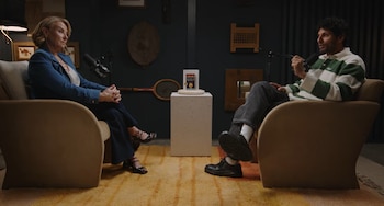 Dos personas, una mujer y un hombre, sentadas en sillones club individuales color beige, se miran con micrófonos frente a ellos en un estudio oscuro