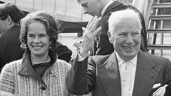 Fotografía en blanco y negro de Charlie Chaplin y Oona O'Neill. Chaplin, con traje, sonríe y saluda con la mano. O'Neill, con collar de perlas, también sonríe