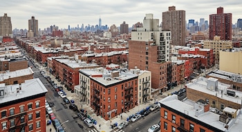 Vista aérea de un barrio en Nueva York con edificios residenciales de ladrillo rojo, calles con coches y el horizonte de Manhattan y sus rascacielos al fondo.
