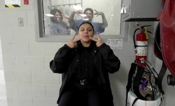 Una mujer vestida de negro realiza gestos con sus manos frente a una ventana, detrás de la cual dos mujeres con ropa azul hacen señas