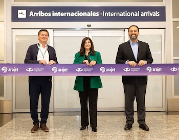 Tres personas, dos hombres y una mujer, sonríen mientras sostienen una cinta morada con logotipos frente a un letrero de "Arribos internacionales" en un aeropuerto
