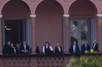 Varias personas, incluyendo hombres en traje y una mujer, observan desde el balcón de un edificio rosado con arcos ornamentados