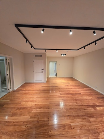 Una habitación vacía con paredes beige, piso de madera pulida, puerta blanca, y un sistema de iluminación moderno con focos negros en el techo