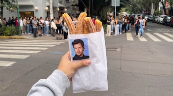 Una mano sostiene una bolsa blanca con churros y un parche de Harry Styles sobre la bolsa; detrás, filas de personas en una calle con un paso de cebra.