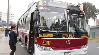 Bus de transporte público negro, rojo y blanco en una calle. Una mujer de uniforme oscuro con cabello rojo está de pie junto al bus