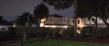 Vista nocturna de varias casas residenciales con tejados oscuros, rodeadas de palmeras y arbustos verdes, iluminadas por luces cálidas contra un cielo oscuro