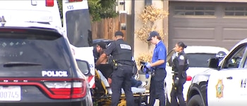 Oficiales de policía y personal médico rodean a una persona en camilla junto a una ambulancia y patrullas frente a una vivienda